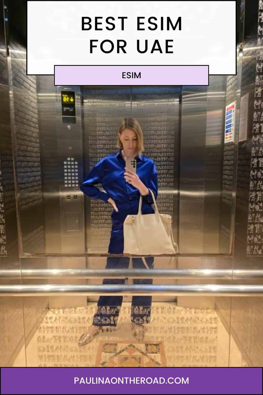 a woman in blue blouse, black pants, and a bag in an elevator taking a picture with a phone